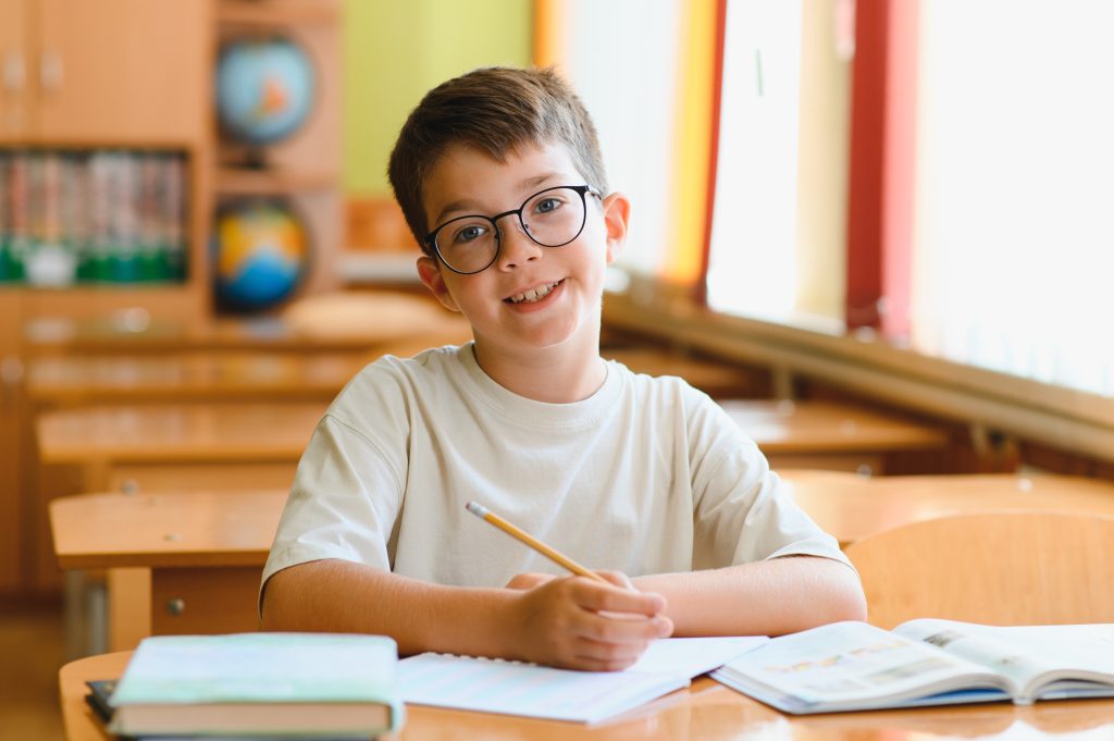 Happy,Young,Boy,Wearing,Eyeglasses,Sitting,At,Desk,In,Classroom